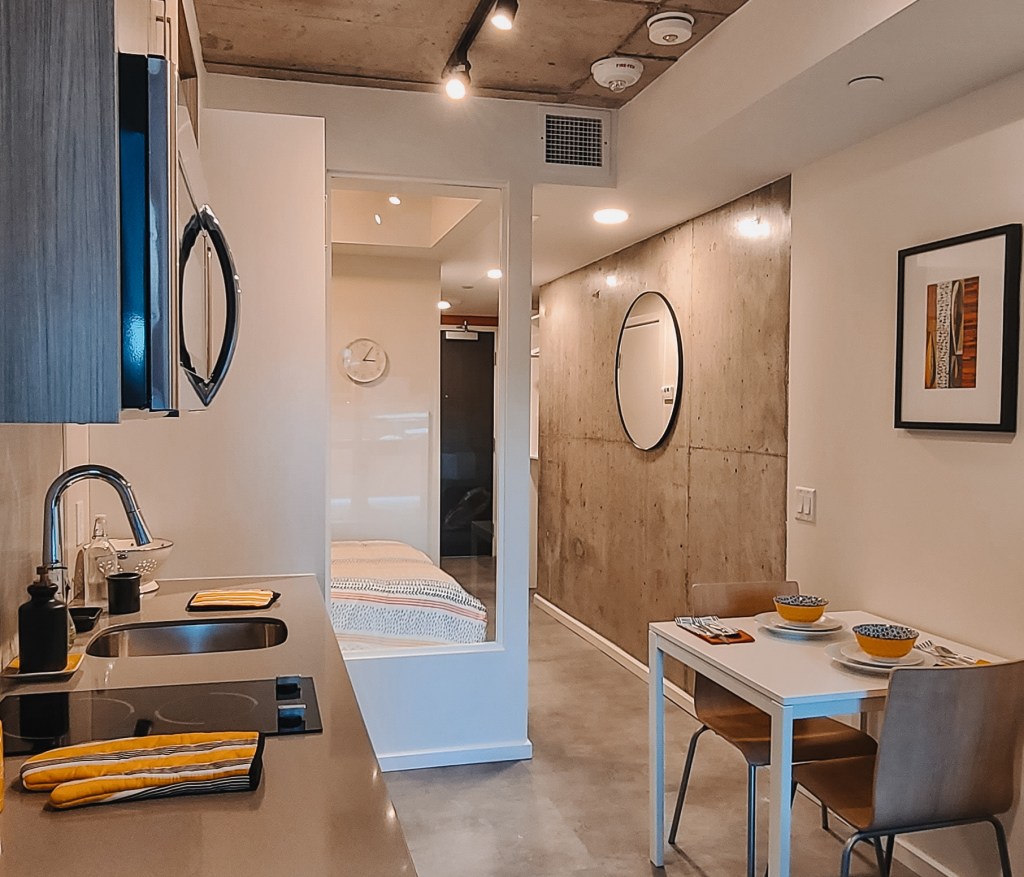 View of Envie suite with kitchen station, dining table, bedroom area and hallway, including design details like mirrors and modern wall art